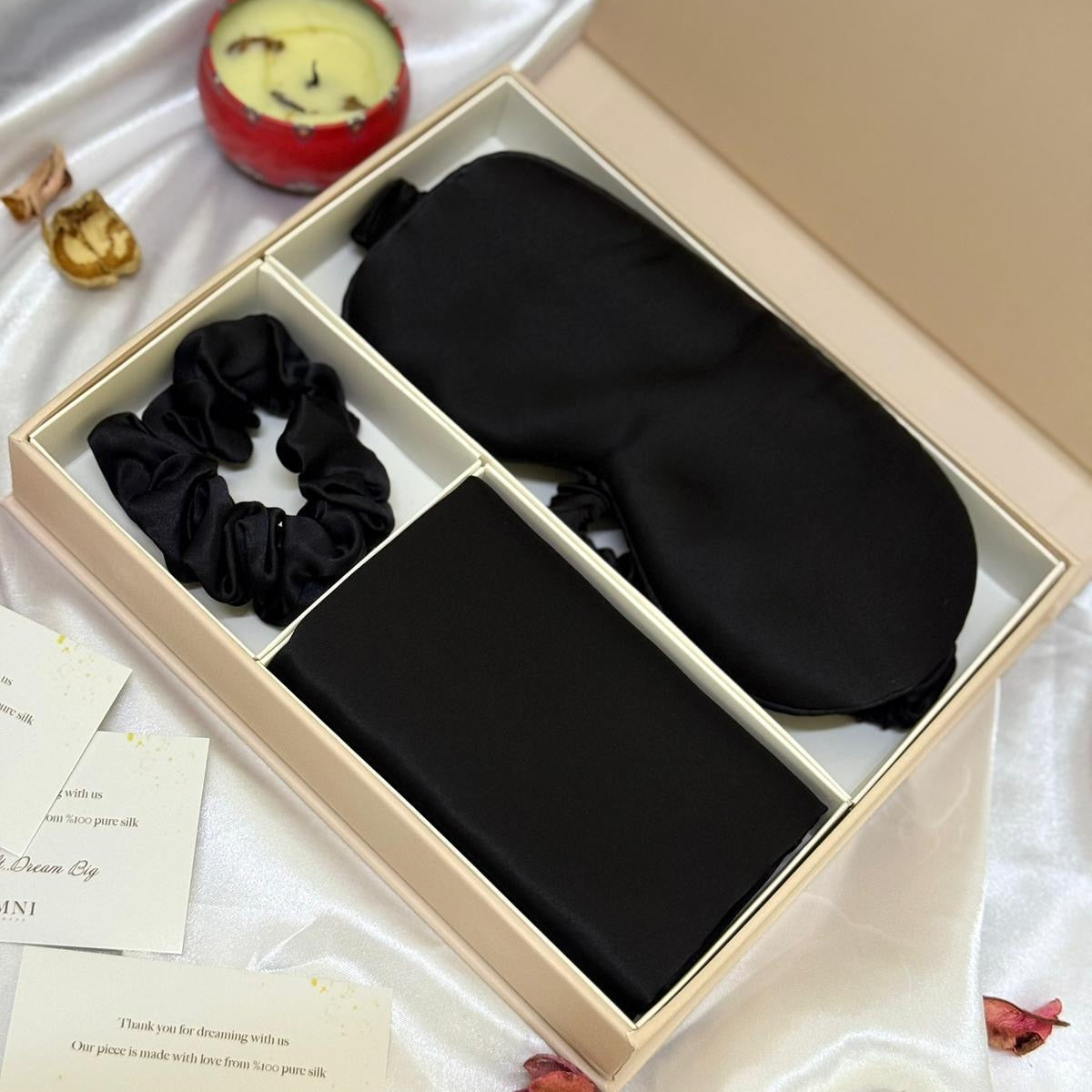 Set of black eye mask, scrunchie, and small pouch in a box with decorative items around.